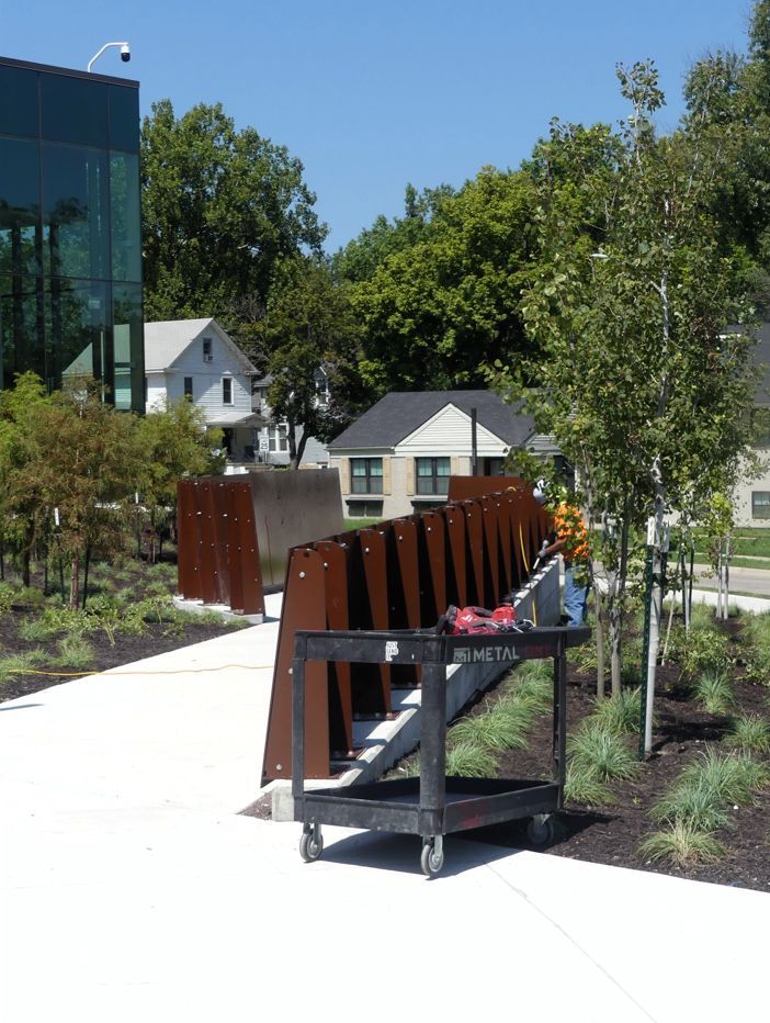 A sidewalk with a rusty metal barrier, a black cart, and a building with glass windows, trees and houses in the background.