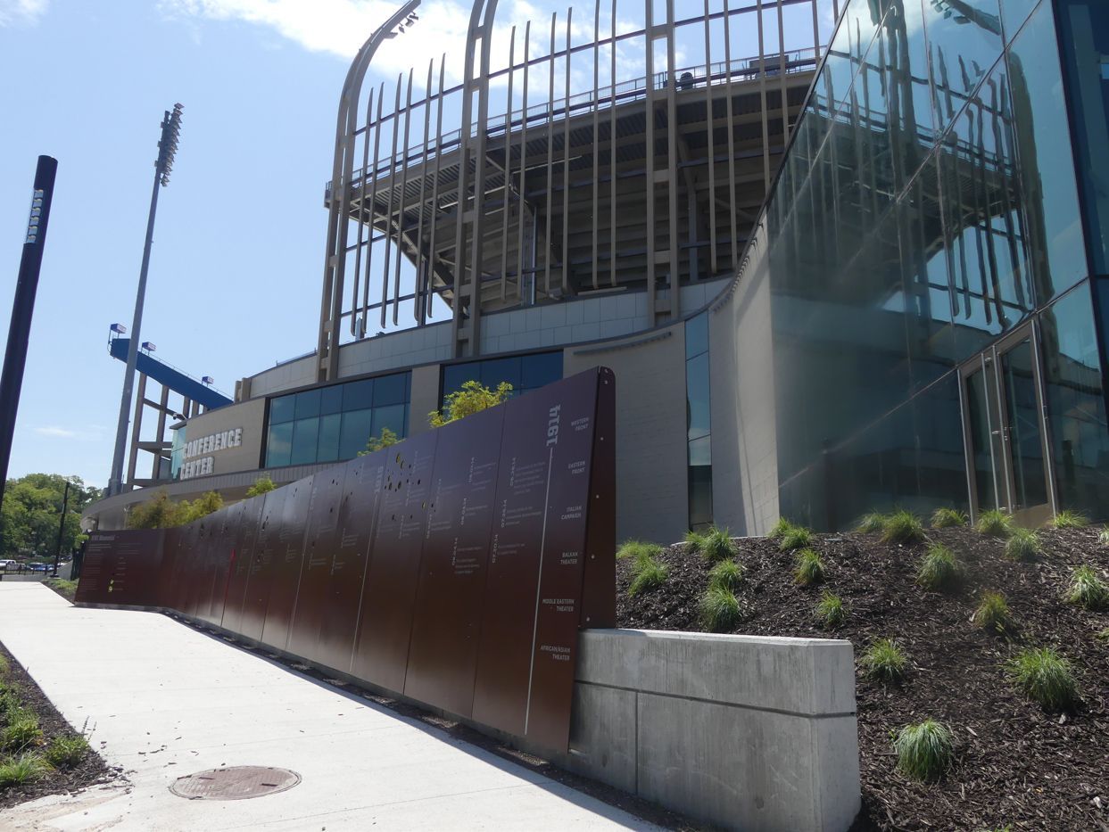 Exterior view of a modern stadium under construction with brown steel walls, green grass, and a blue lift.
