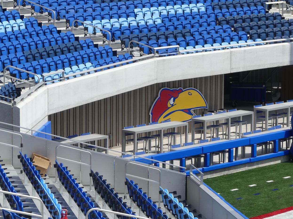 A stadium with blue seating, a large Jayhawk logo, and outdoor tables.
