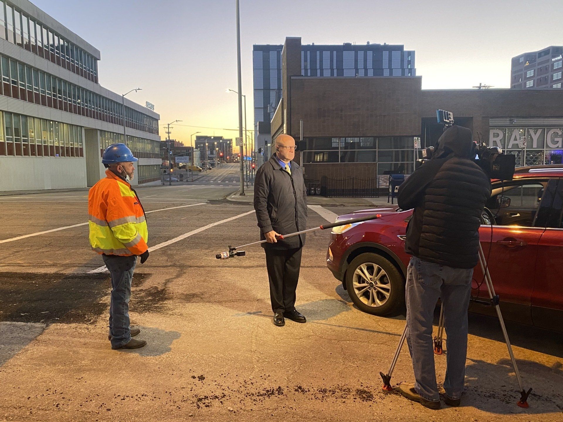 A man is standing in front of a red car talking to a cameraman.
