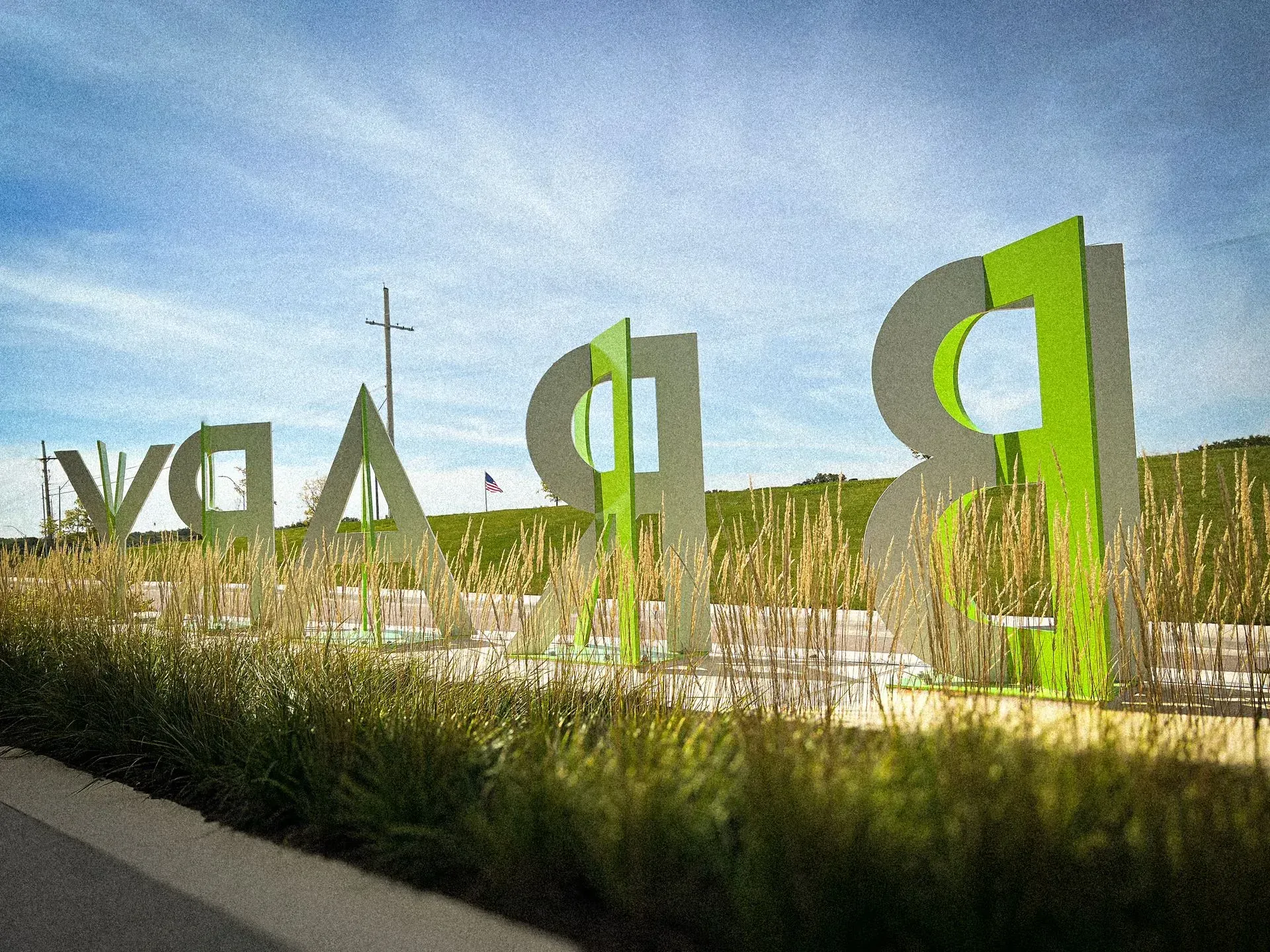 Large metal sign in green and silver against tall grass and a blue sky.