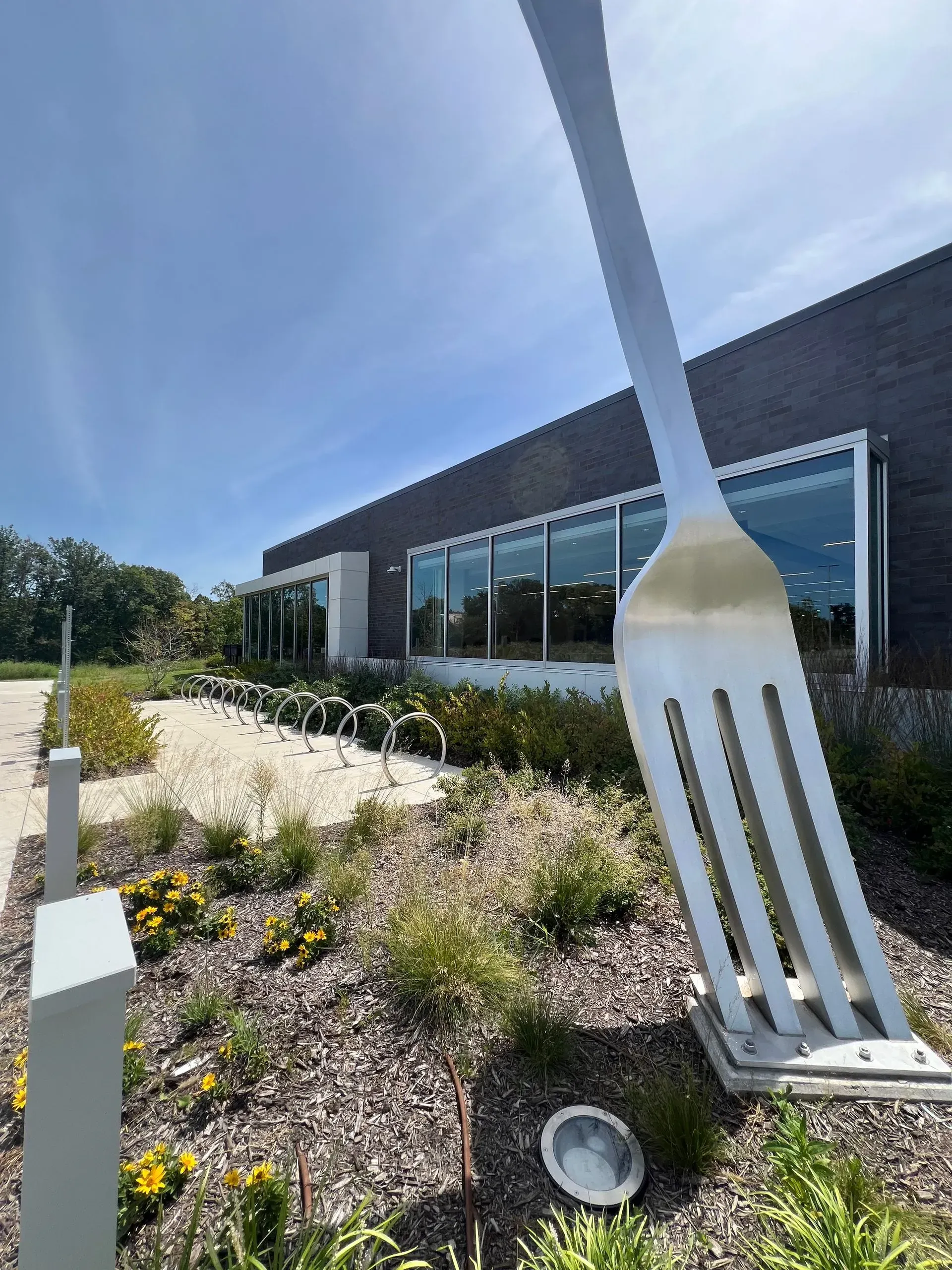 Giant fork sculpture in a garden setting, in front of a modern building with a blue sky background.
