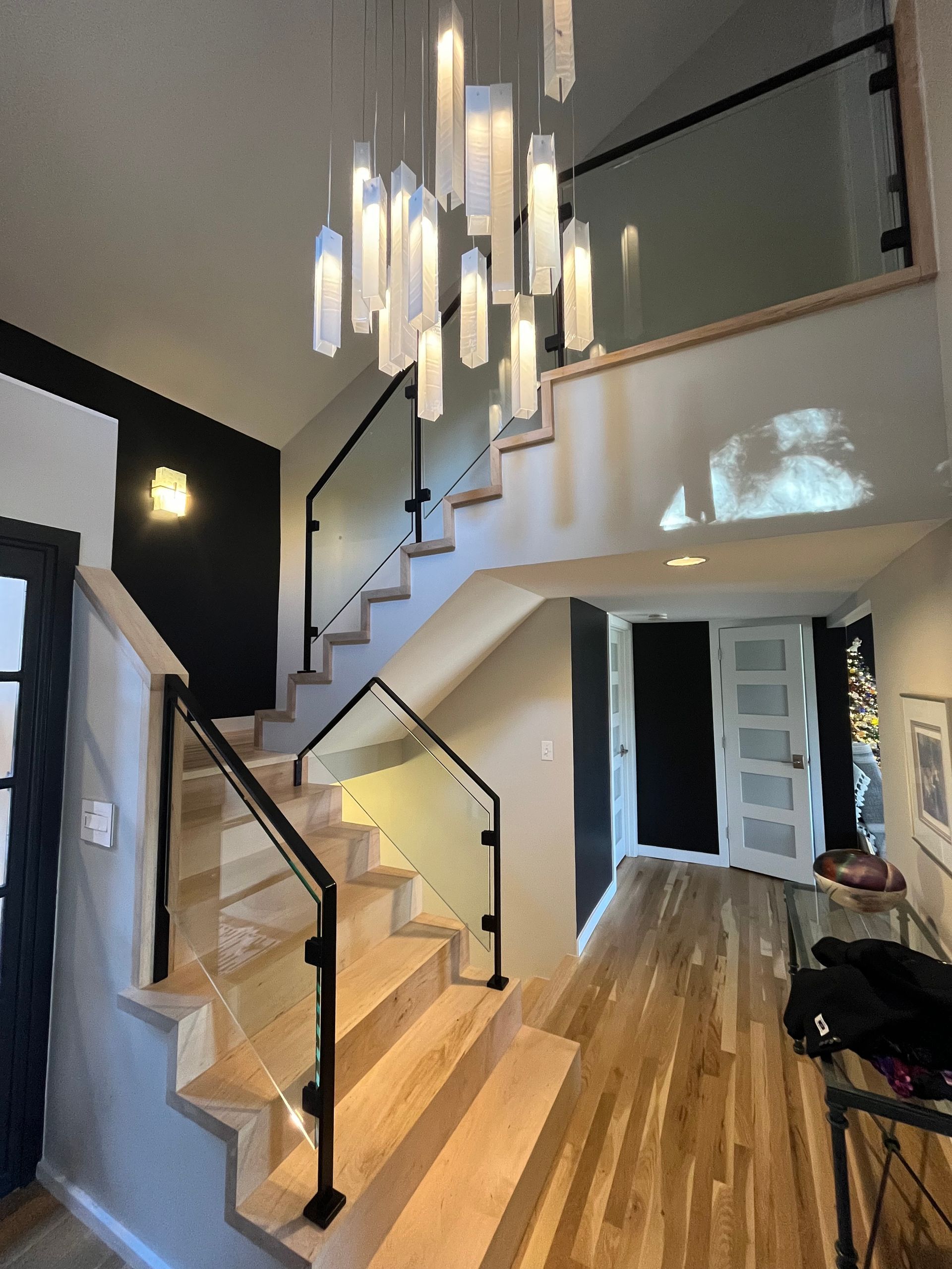 Staircase with glass railings, light wood steps, and a modern chandelier in a bright entry hall.