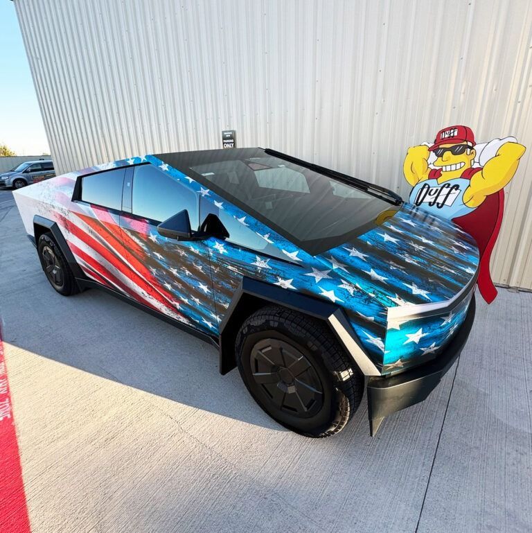 Cybertruck wrapped in American flag colors parked beside a white wall and cartoon sign