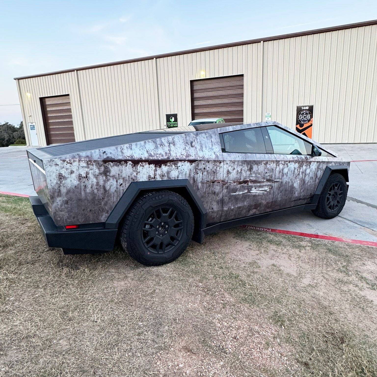 Rusty, armored Tesla Cybertruck parked outside a warehouse on a grassy lot.