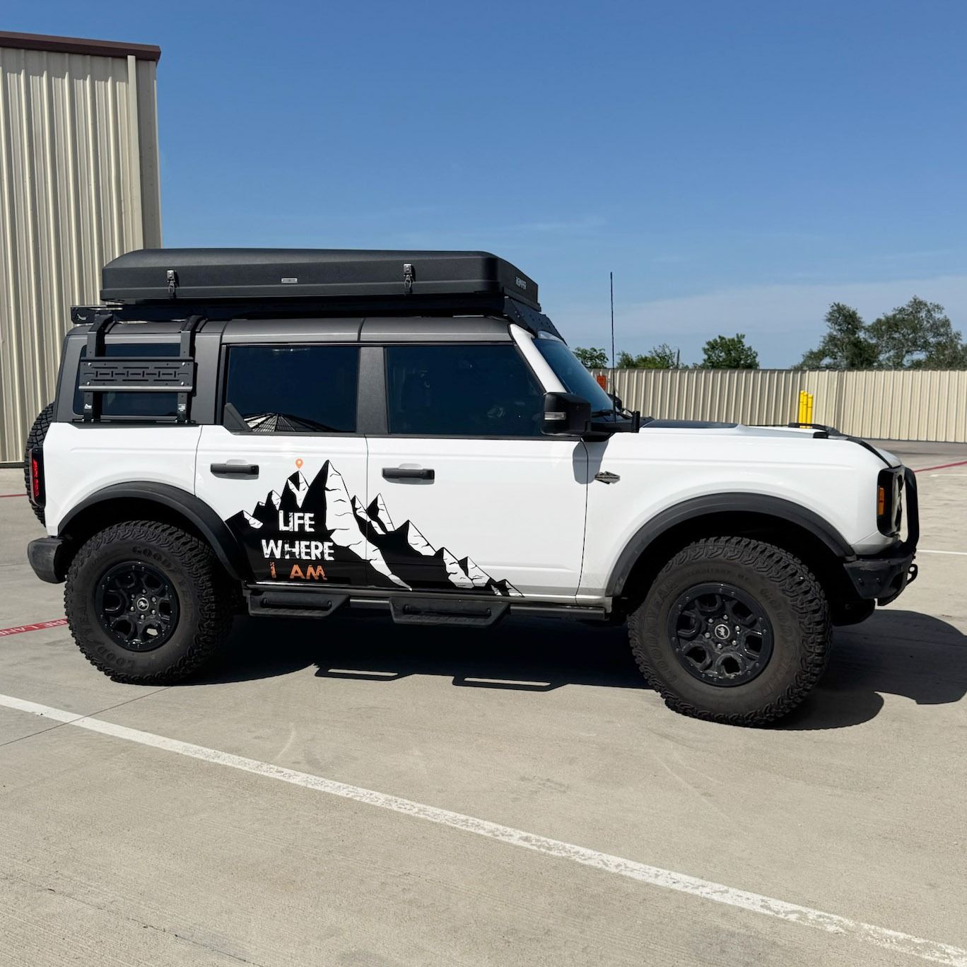 White off-road SUV with black roof tent and mountain decal parked in a lot under a clear blue sky