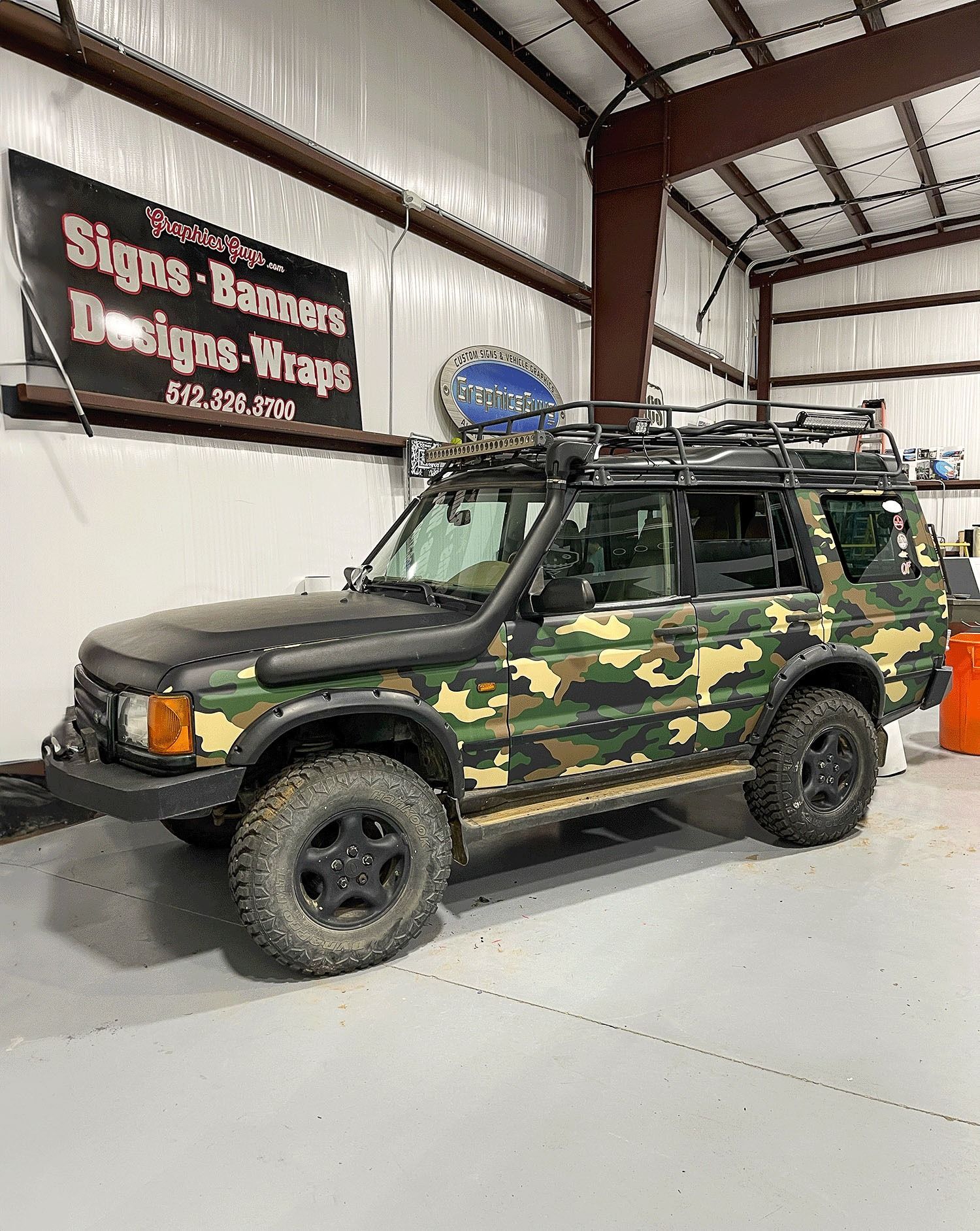 Camouflaged off-road SUV in a warehouse garage with roof rack and muddy tires