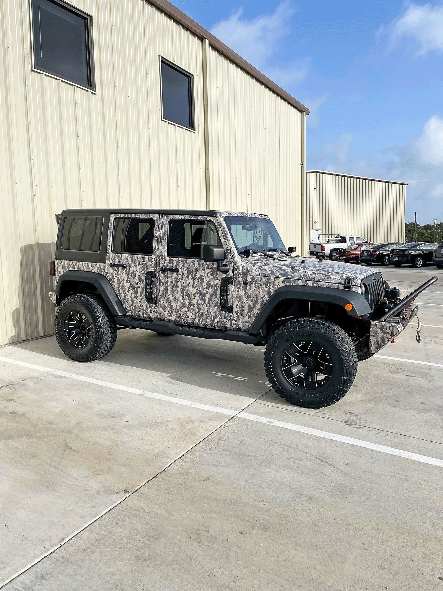 Camouflage Jeep Wrangler parked beside a beige industrial building on a sunny day.