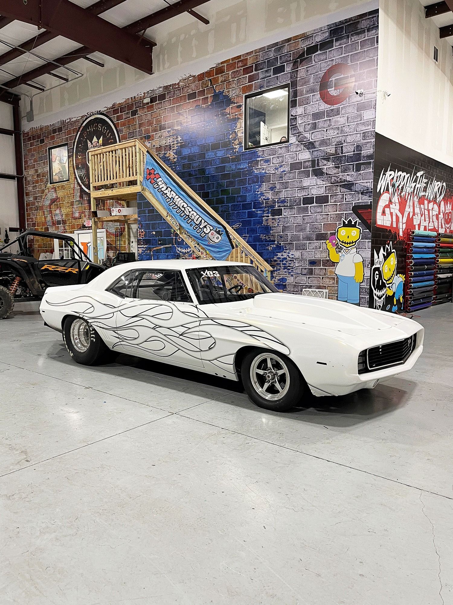 White classic muscle car with black flame graphics in a graffiti-covered garage.