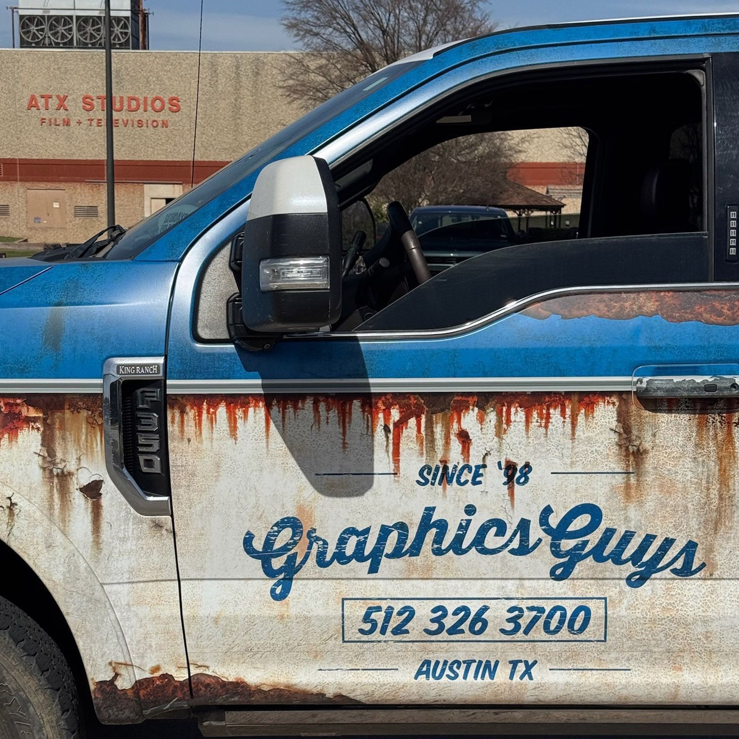 Blue work truck with “Graphics Guys” door logo and phone number, parked by an ATX Studios building.