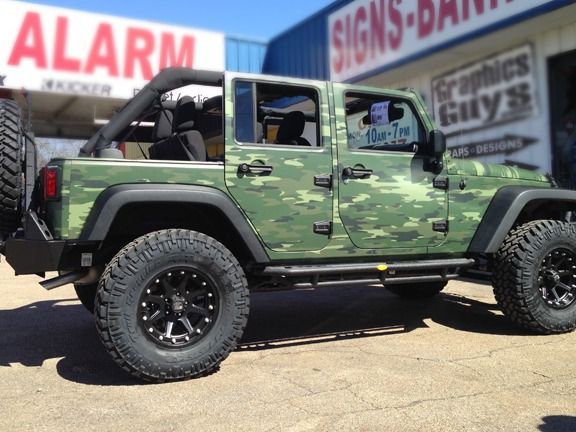 Green Jeep Wrangler on black off-road tires parked outside a sign shop