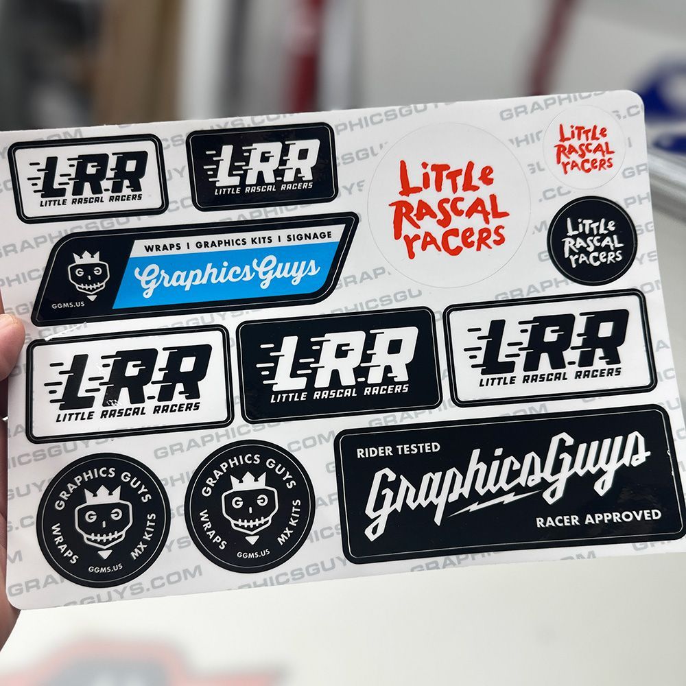 Hand holding a sheet of black, white, red, and blue racing decals and stickers, including “Graphics Guys” and “LRR”
