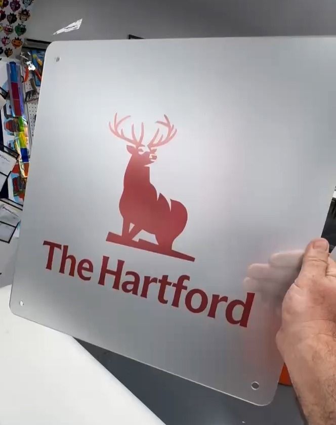 Metal sign with red Hartford deer logo and “The Hartford” text, held by a hand