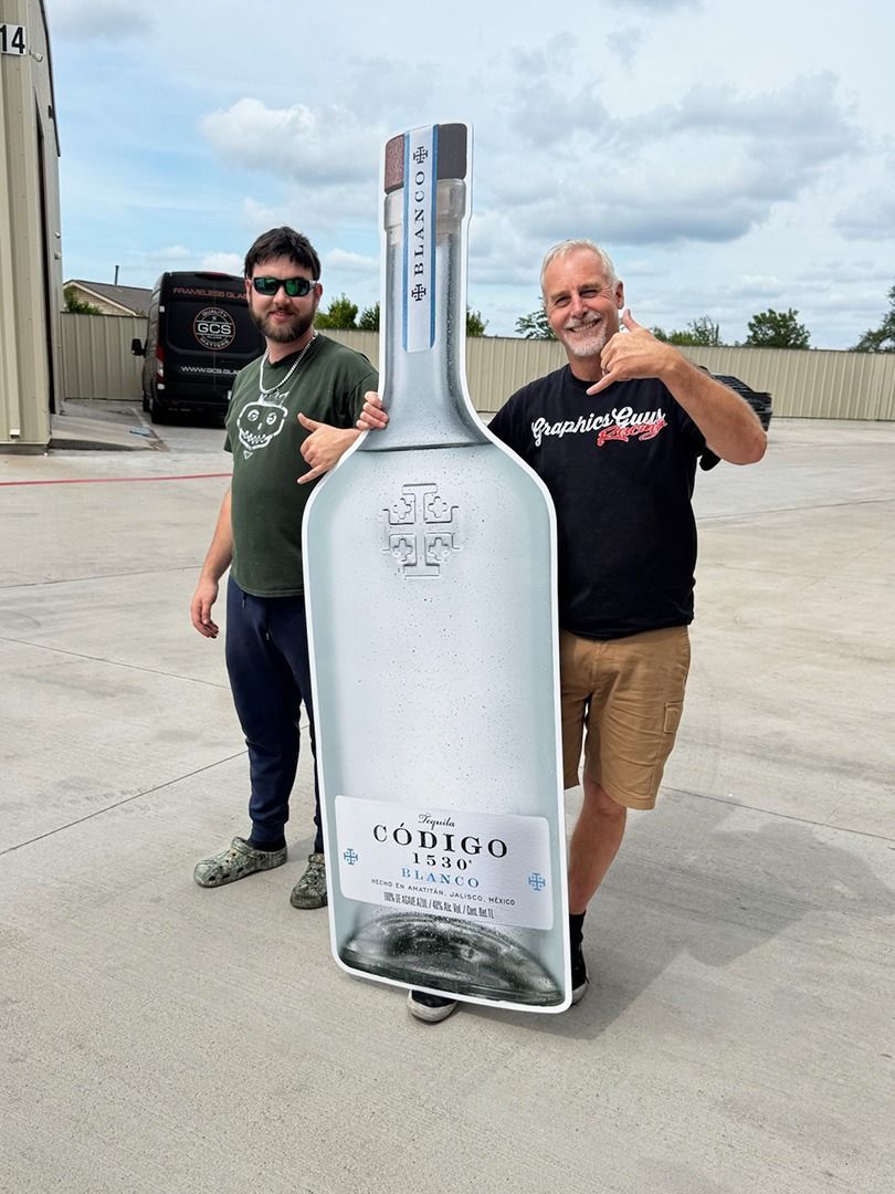 Two men stand beside a giant cutout bottle prop in an outdoor parking lot.