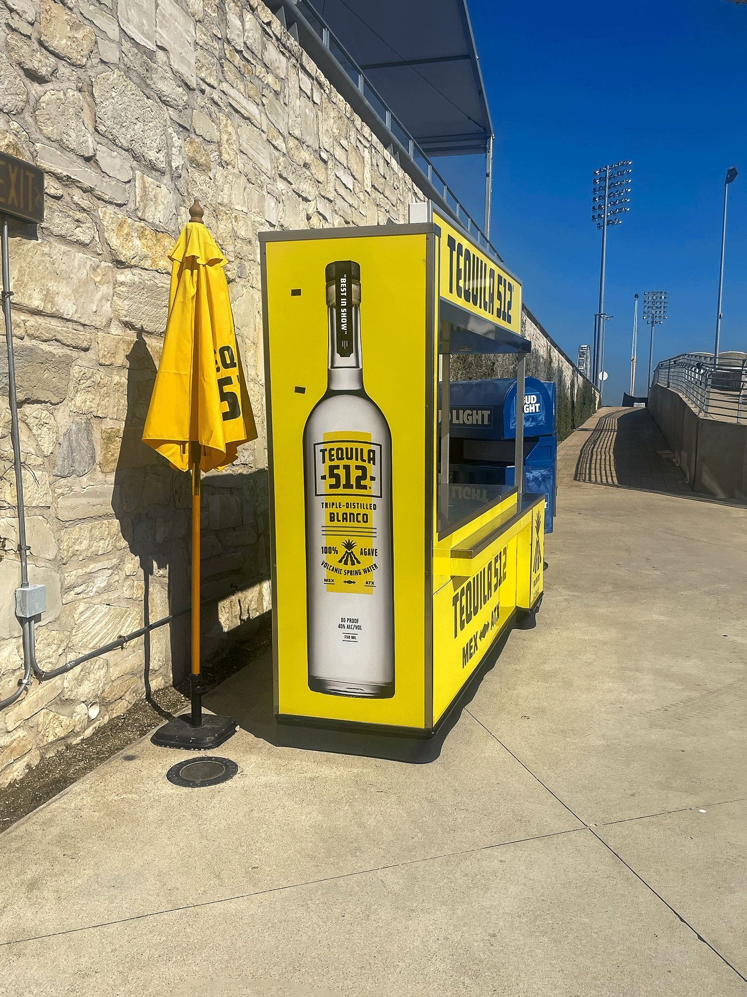 Yellow newspaper vending box and umbrella by a stone wall on a sunny sidewalk.