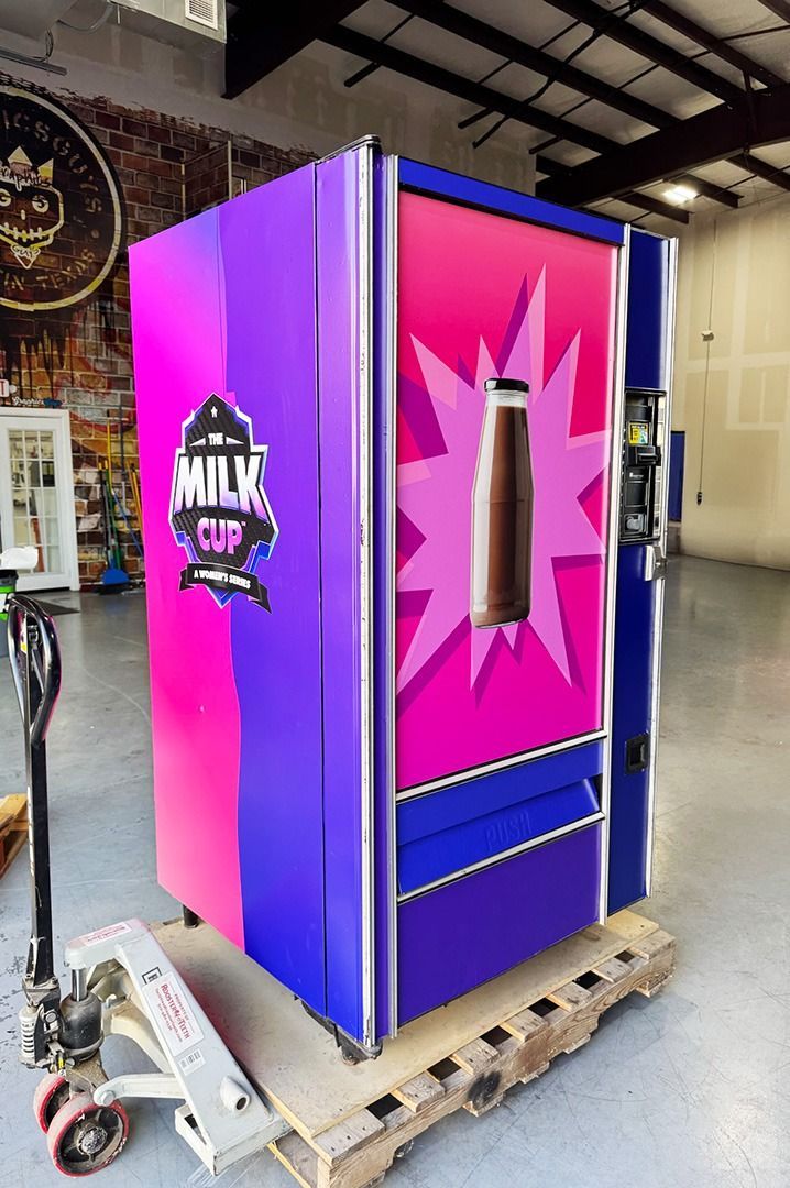 Bright pink and purple arcade-style kiosk with a central cutout, sitting on a wooden pallet in a warehouse.