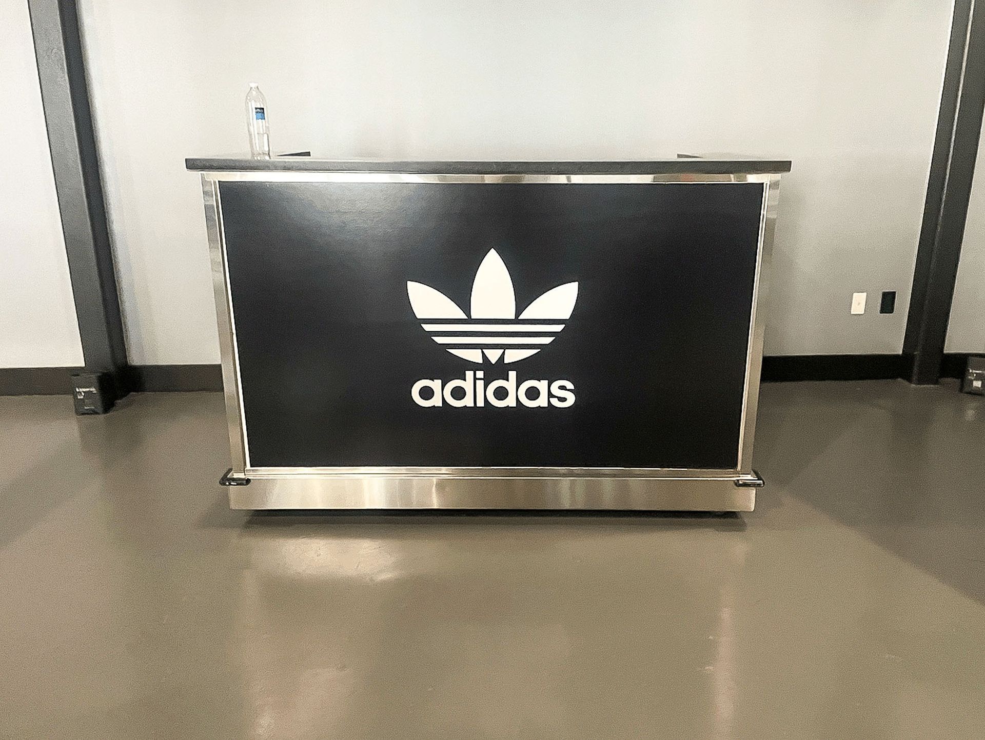 Adidas-branded black counter with white logo in an empty room, against a gray wall.
