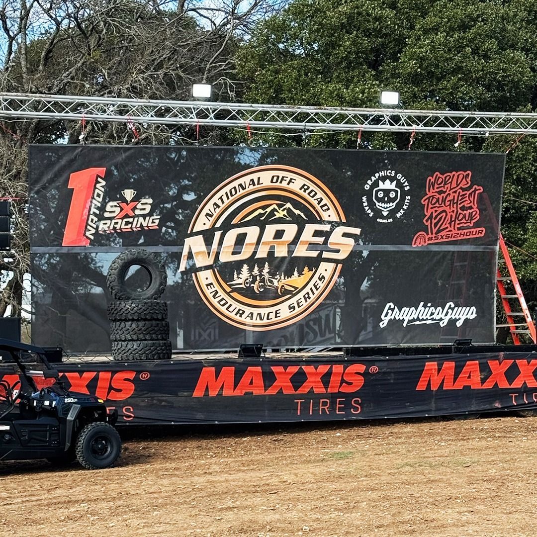 Black off-road truck before sponsor banners with NORS logo and Maxxis branding at a dirt track