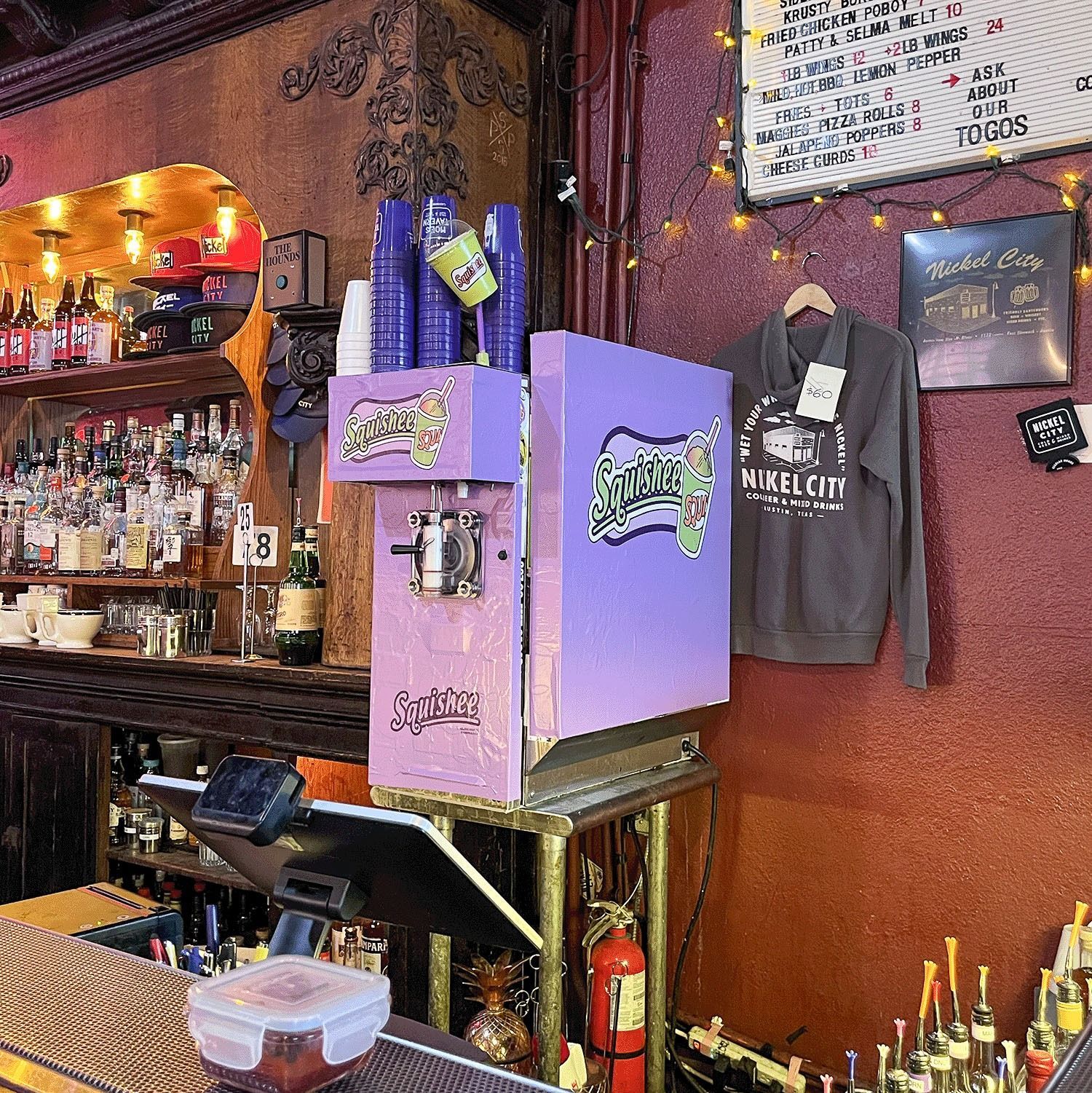Bar shelf with liquor bottles, purple cash box, hanging T-shirt, and drink menus in a cozy bar interior