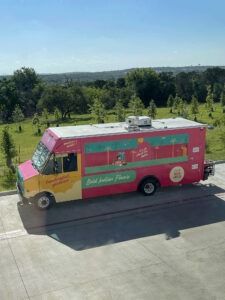 Pink food truck parked on a road beside green trees and open countryside under a clear sky