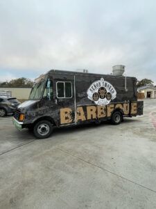 Black food truck with large “BARE” lettering parked on a concrete lot under a cloudy sky