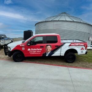 State Farm branded red-and-white pickup truck parked beside a grain silo under a blue sky.