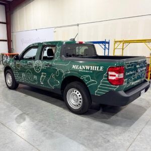 Green pickup truck with white graphics and “MEANWHILE” branding parked indoors in a warehouse.