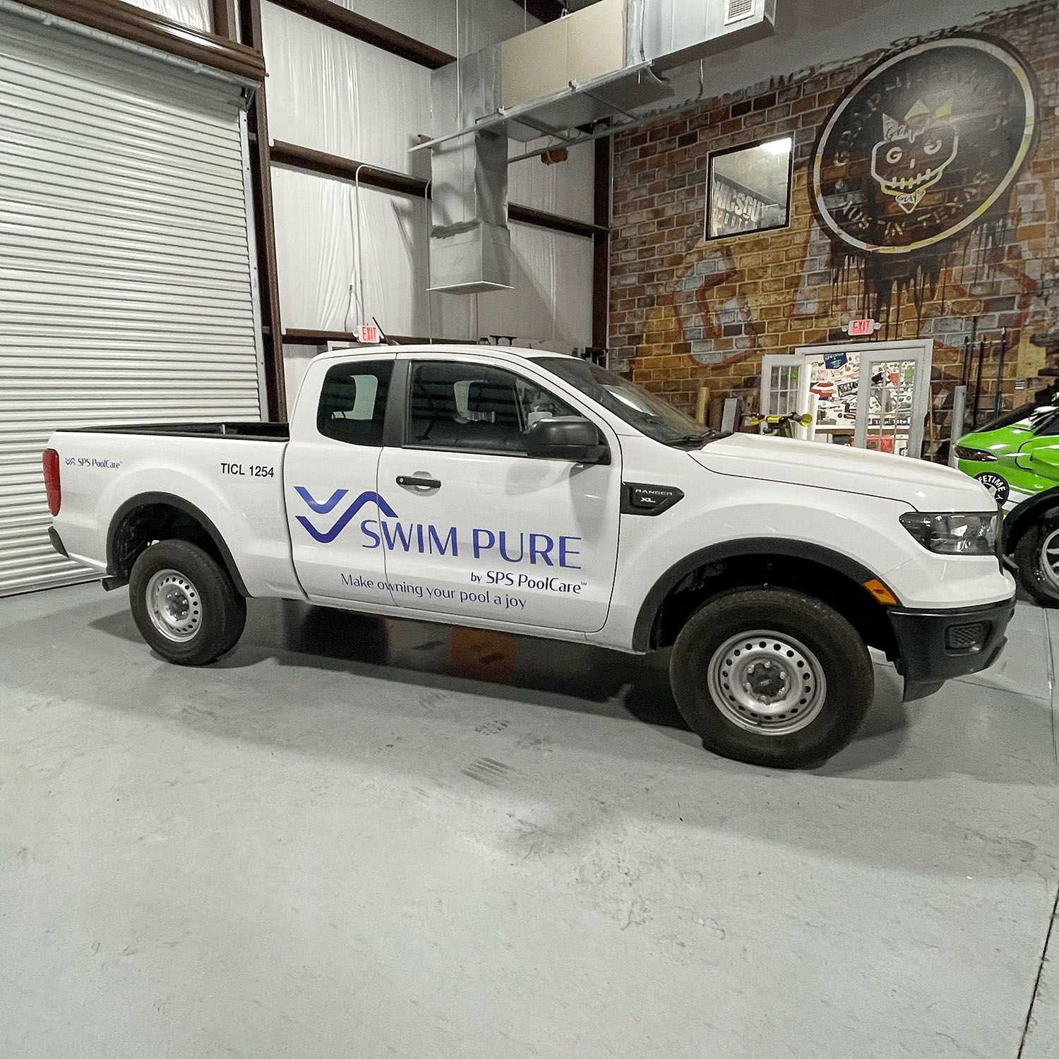 White SWIM PURE pickup truck parked indoors beside a green vehicle in a warehouse garage.