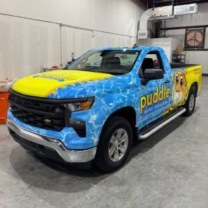 Blue and yellow Puddle pickup truck wrapped with water splash graphics, parked indoors.
