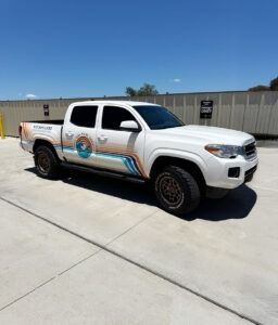 White pickup truck with logo decals parked on a sunny concrete lot beside a fence
