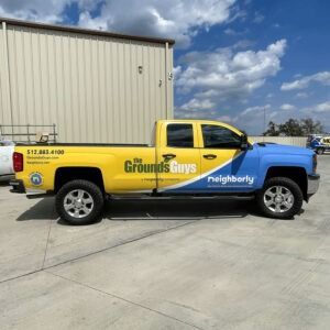 Yellow-and-blue Grounds Guys pickup truck parked outside a building under a partly cloudy sky