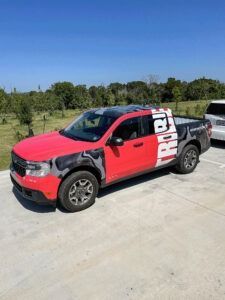 Red pickup truck with black and white graphics parked on a concrete lot beside grassy trees under a blue sky