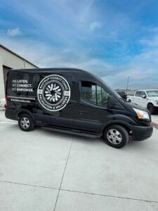 Black delivery van with white logo parked outdoors on a concrete lot under a blue sky.