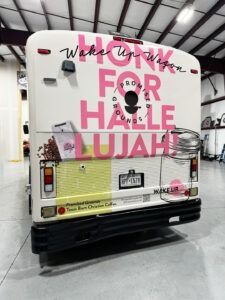 White bus in a warehouse with large pink “FOR SALE” lettering on the rear, plus other decals and a black license plate.