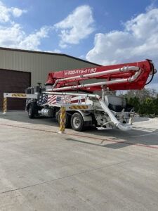 Red concrete pump truck parked outside a warehouse under a blue sky