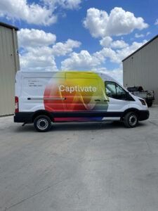 White delivery van with colorful “Captivate” wrap parked between warehouse buildings under a cloudy sky