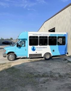 Light blue and white shuttle bus parked beside a beige building on a gravel lot.