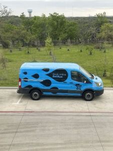 Blue service van with black flame decals parked on a concrete road beside a grassy field and trees.