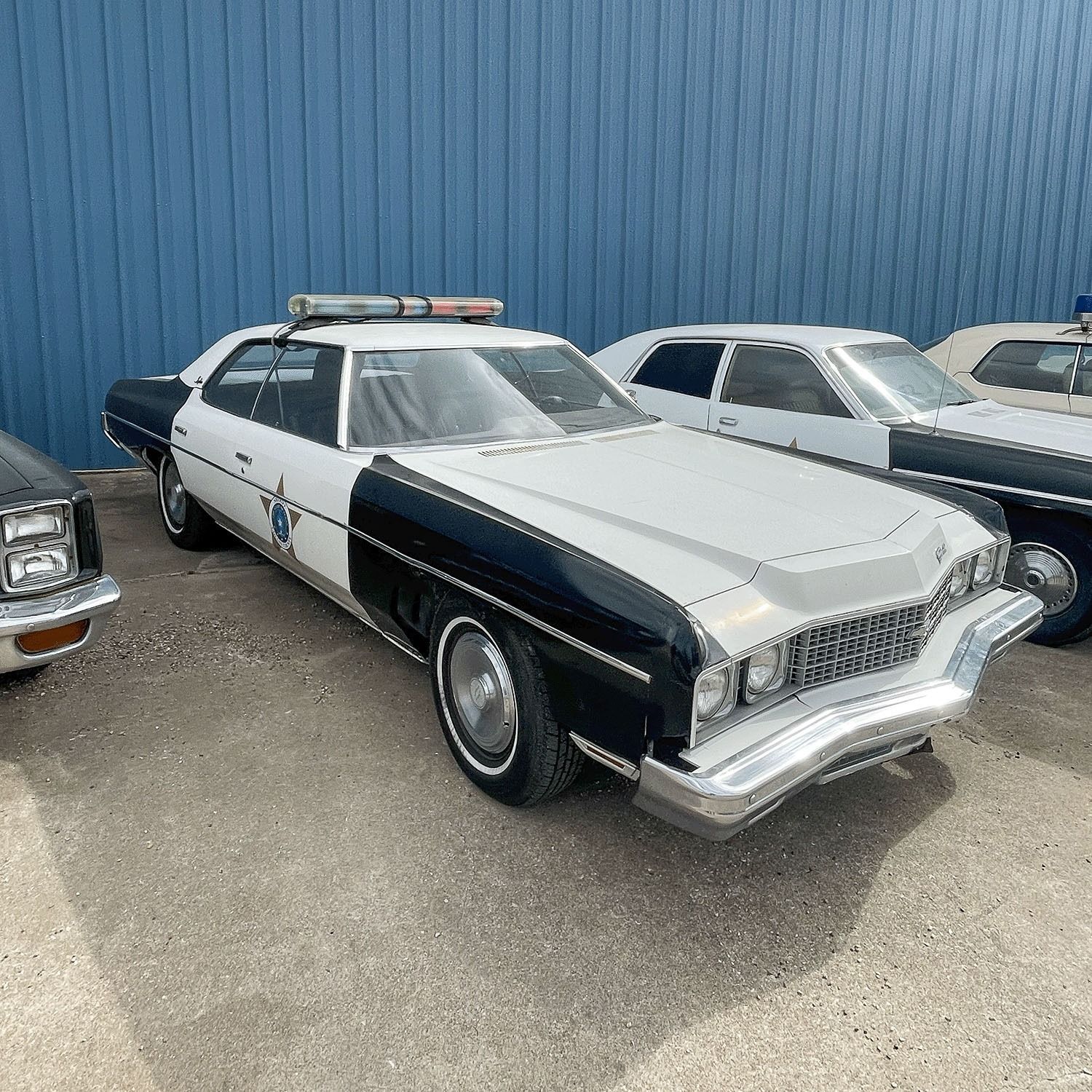 Black-and-white vintage police car parked beside another classic car against a blue wall