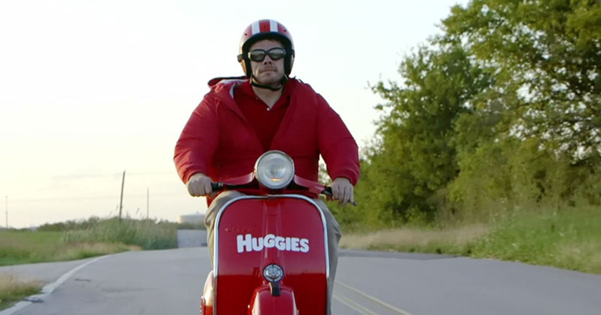 Person in a red jacket riding a red scooter on a rural road, with trees and grass alongside.