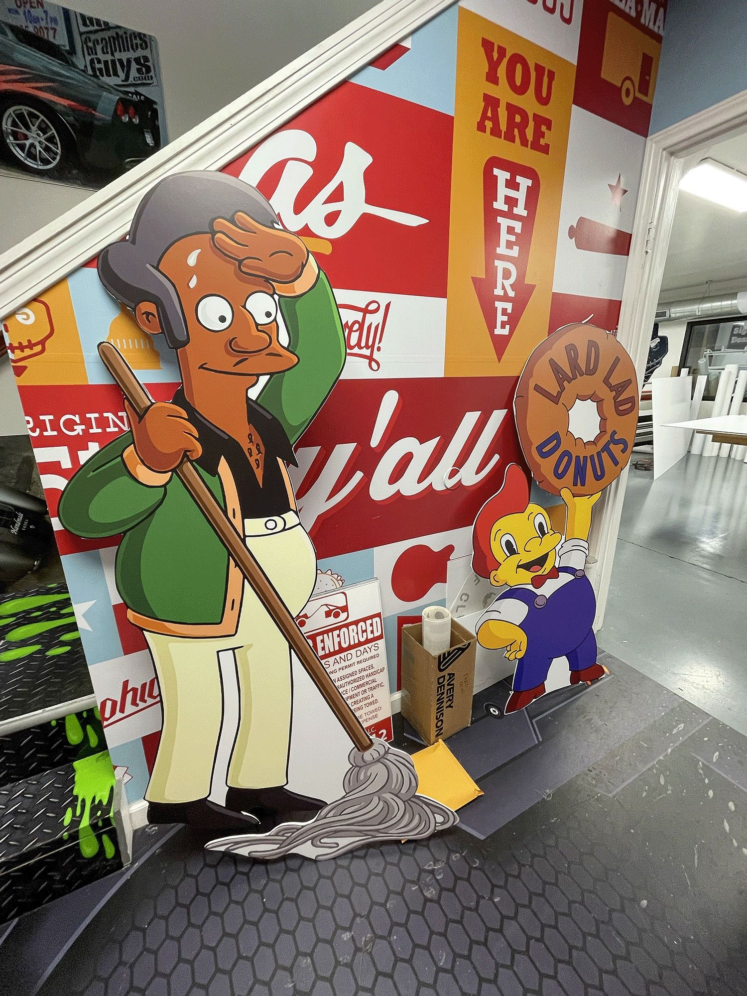 Cartoon cutouts of people on a mall staircase beside a red wall and a “You Are Here” sign