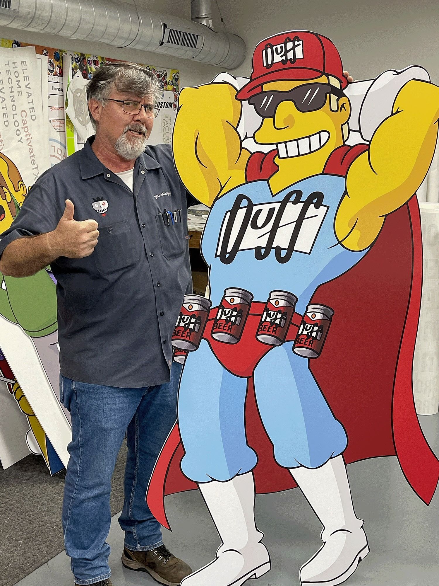 Man standing beside a large yellow cartoon cutout in a workshop, giving a thumbs-up.