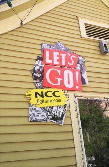 Red “LET’S GO!” sign above yellow “NCC digital media” poster on a house wall