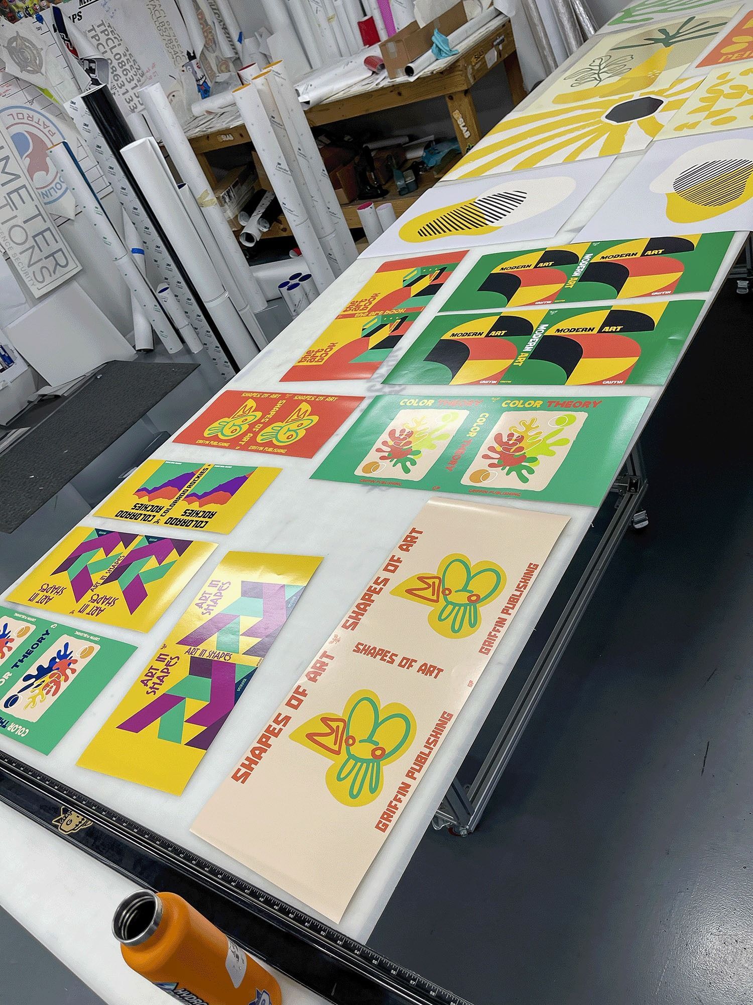 Table of colorful abstract prints displayed on a print shop worktable, with an orange drink bottle in the foreground