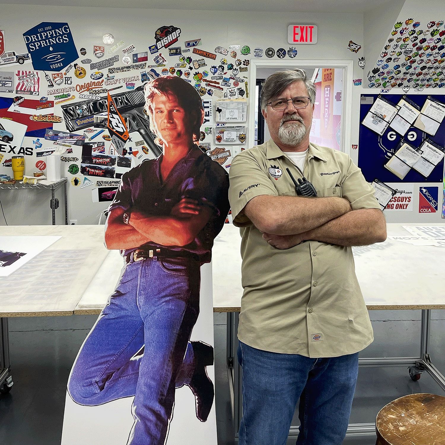 Life-size cutout beside a man standing with arms crossed in a sticker-covered room.