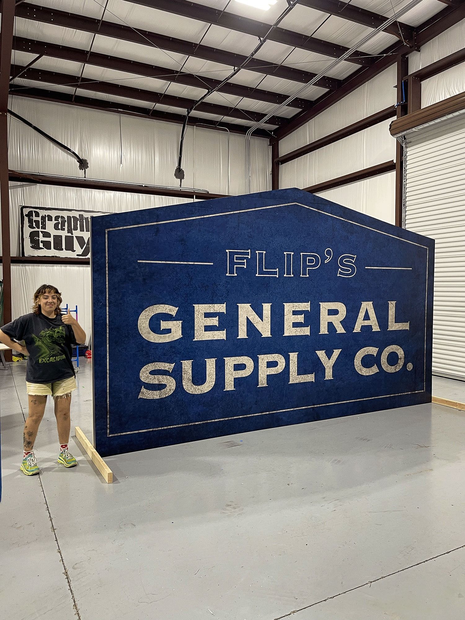 Woman standing beside large blue “General Supply Co.” sign in an industrial warehouse