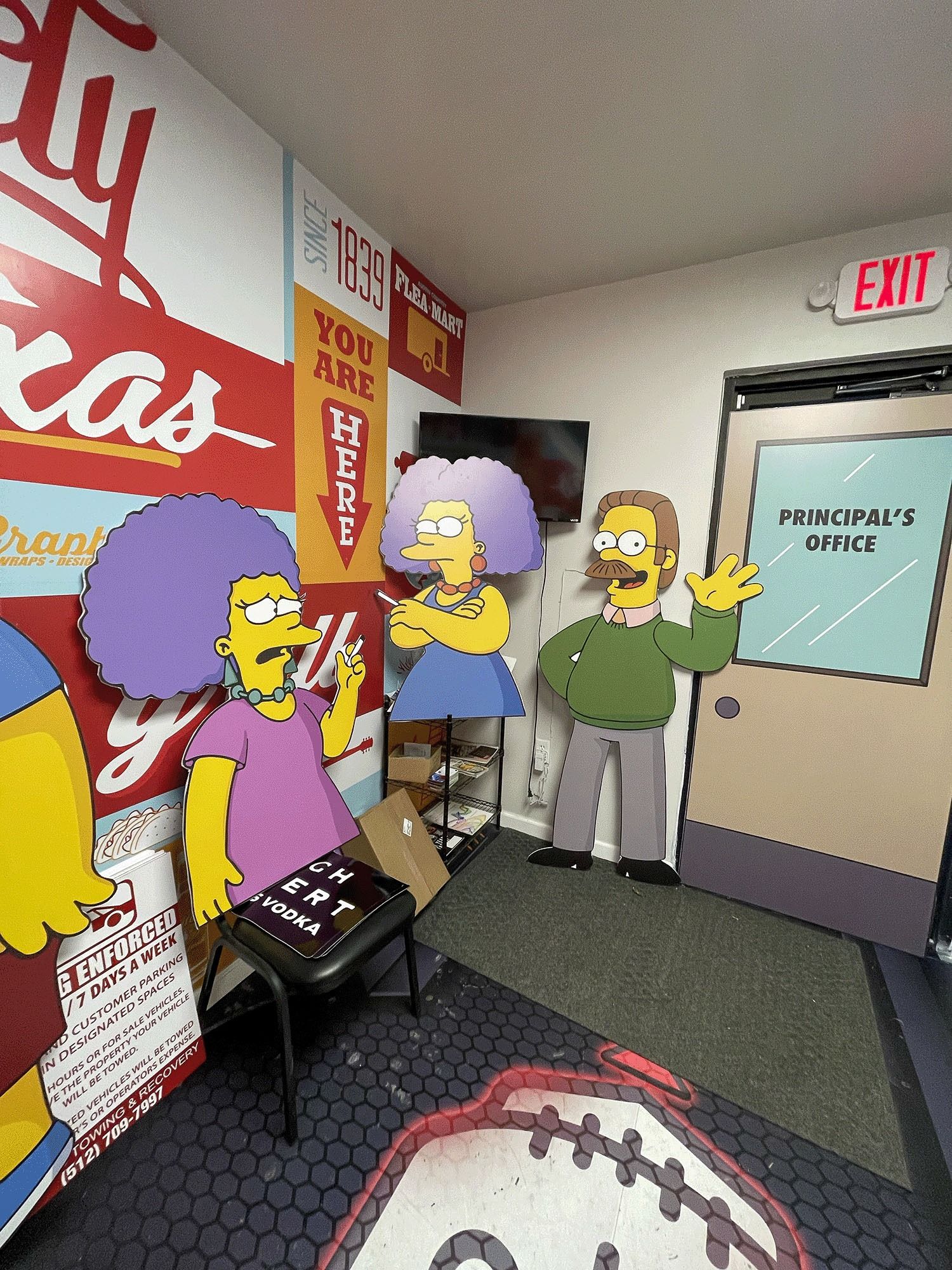 Simpsons characters in a narrow office hallway with a “Principal’s Office” door and exit sign.