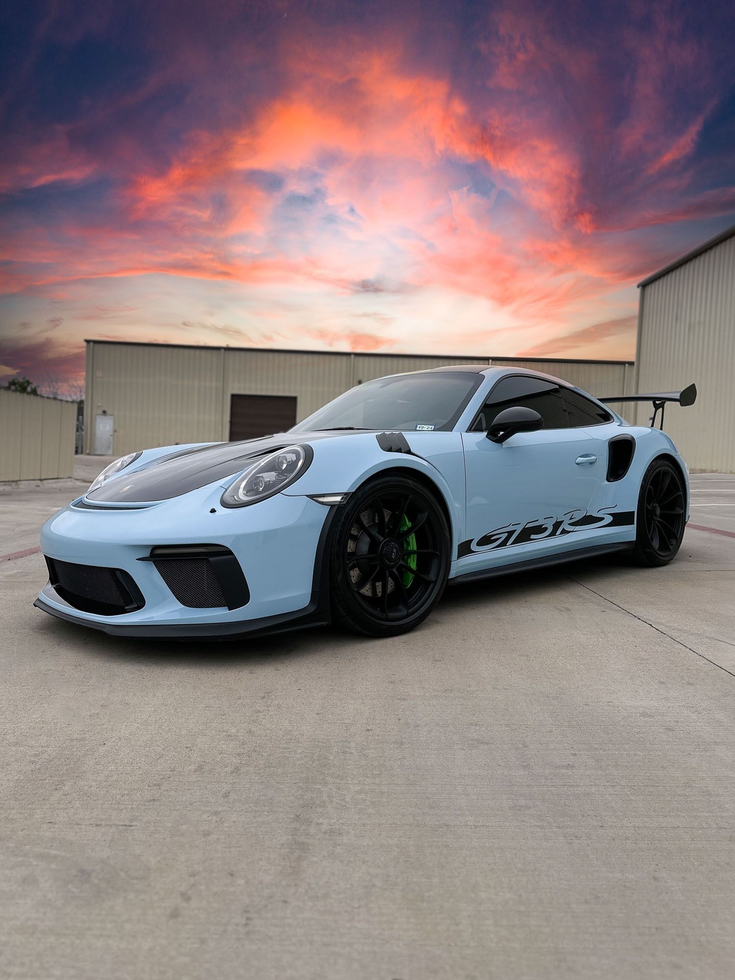 Light blue sports car parked in an industrial lot at sunset with a colorful sky