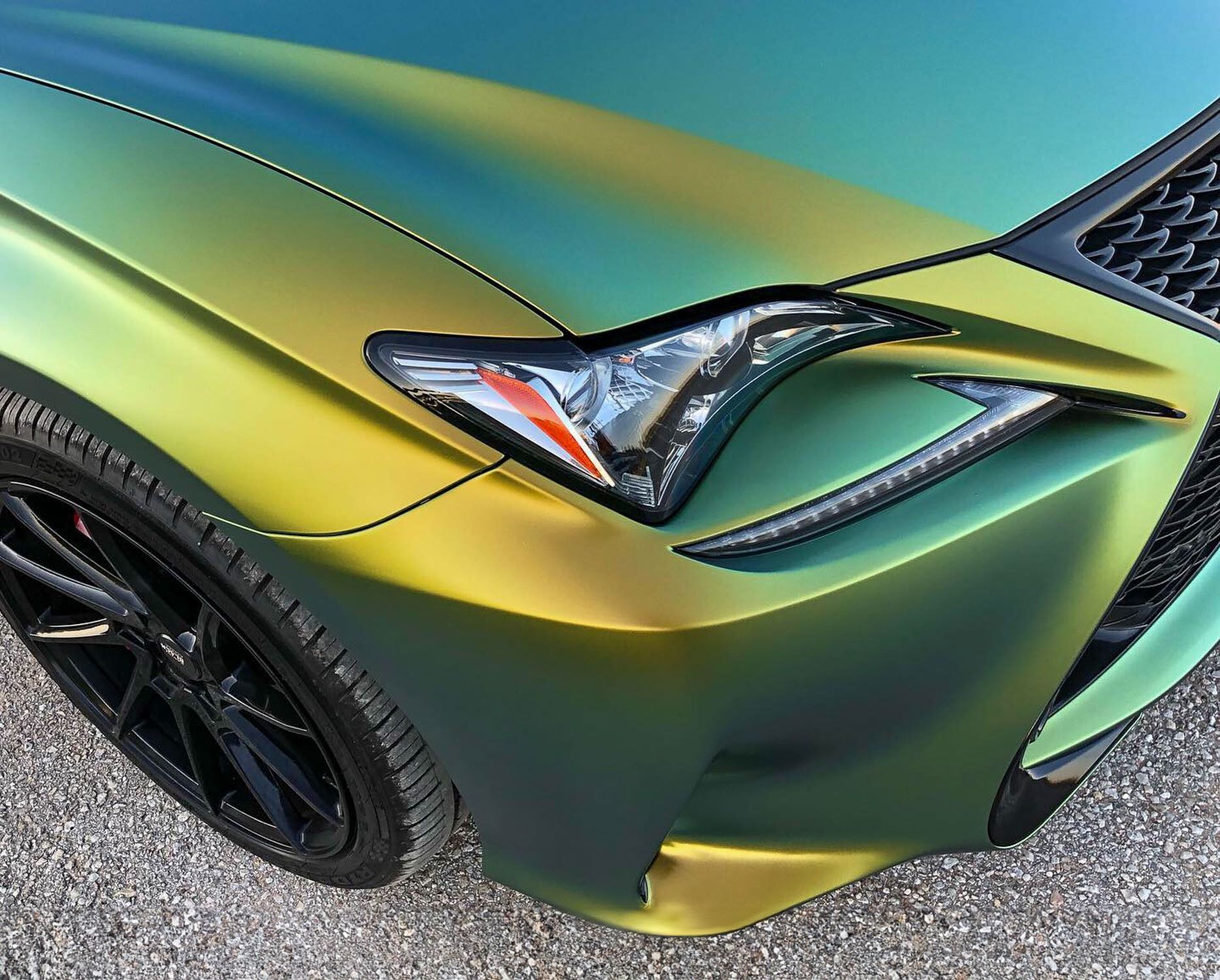 Close-up of a matte green sports car front fender, headlight, and black wheel on pavement.