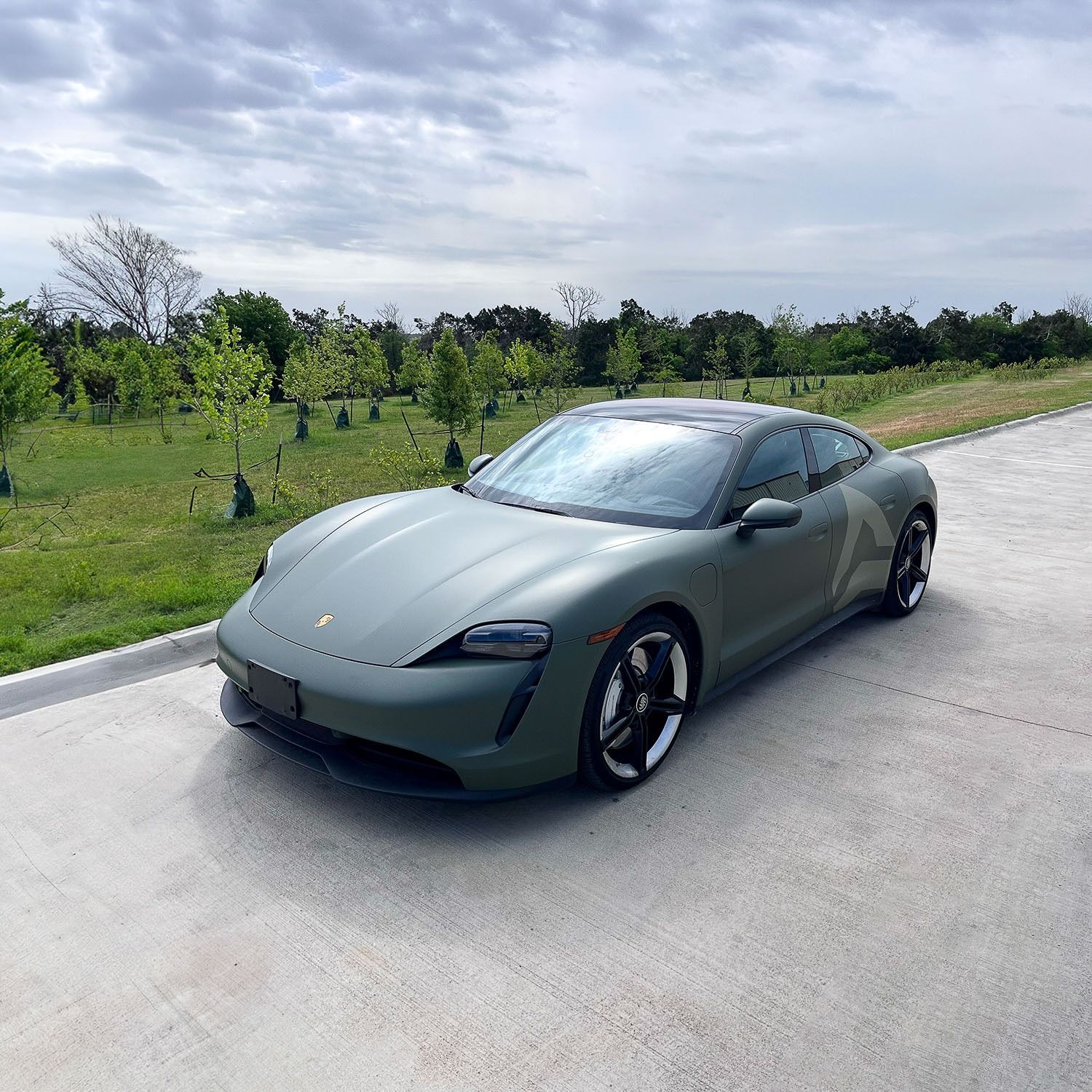 Matte gray sports car parked on a paved road beside a grassy field under a cloudy sky